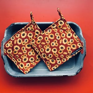 Könnte beinhalten: Zwei quadratische Topflappen mit Sonnenblumenmuster liegen in einer rechteckigen Metallschale. Die Topflappen haben einen roten Hintergrund mit gelben Sonnenblumen. Die Schale ist grau mit rotem Hintergrund.