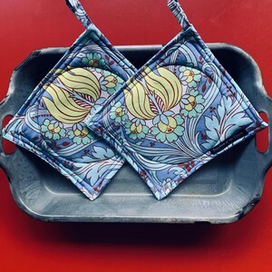 May include: Two square pot holders with a floral pattern in shades of blue, green, and yellow. The pot holders have a quilted design and a loop for hanging. They are placed in a grey metal tray against a red background.