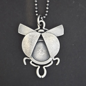 May include: A silver-toned pendant necklace featuring a stylized insect design. The pendant has a central circular element with radiating lines, flanked by two curved, patterned wings. The necklace chain is a dark, beaded style.