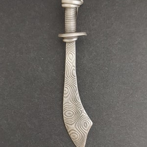 Puede incluir: Un collar con un colgante de plata que representa una espada con un diseño detallado y estampado. El colgante está unido a una cadena de plata.