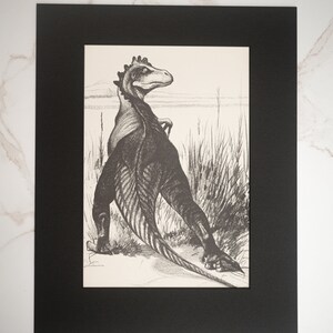 May include: A black and white drawing of a dinosaur standing in a field of tall grass. The dinosaur is facing away from the viewer and has its head turned to the side. The drawing is framed in a black mat.