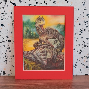 May include: A framed watercolor print featuring two dinosaurs in a prehistoric scene. The dinosaurs are depicted in shades of brown, gray, and green, set against a backdrop of trees and a yellow sky. The print is matted and framed in a red frame.