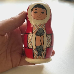 Può includere: Una bambola russa in legno dipinta in rosso, bianco e beige. La bambola raffigura una figura con un cappuccio bordato di bianco che tiene un pesce grigio su uno sfondo a motivi geometrici. Il design presenta contorni neri e una finitura liscia e lucida.