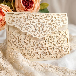 Op de afbeelding: Ivoor lasergesneden uitnodiging met bloemen- en krulwerkontwerp. De ingewikkelde details creëren een delicate en elegante uitstraling. De uitnodiging is open en onthult een glimp van de gedrukte tekst binnenin. Perfect voor bruiloften of speciale evenementen.