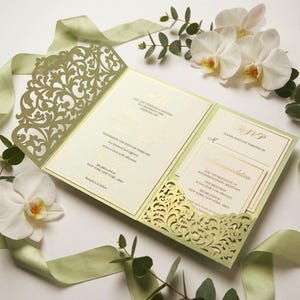 Könnte beinhalten: Eine hellgrüne Hochzeitseinladung mit aufwendigen Laser-Cut-Details. Die Einladung ist geöffnet und zeigt goldenen Text, eine RSVP-Karte und Informationen zur Unterkunft. Weiße Orchideen und grünes Band ergänzen das elegante Design.