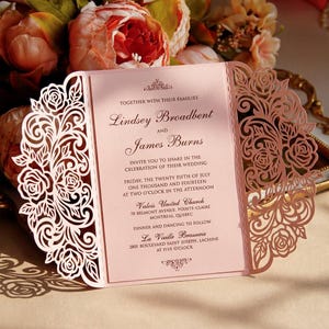 May include: A pink wedding invitation with detailed laser-cut floral patterns. The invitation features the names "Lindsey Broadbent" and "James Burns", and wedding information. The invitation is displayed in front of a floral arrangement.