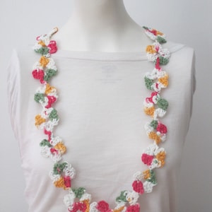 May include: A crocheted flower lei necklace in shades of green, white, pink, and yellow. The lei is made of individual crocheted flowers that are linked together.