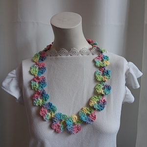 Puede incluir: Un collar de ganchillo colorido con diseños en forma de flor en tonos pastel de rosa, azul, amarillo y verde. El collar se exhibe en un maniquí blanco, mostrando la artesanía hecha a mano y los colores vibrantes.