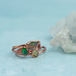 May include: A stack of three thin copper rings, each featuring a small, raw, irregularly shaped gemstone. The stones display iridescent green and translucent white hues, set within simple metal bands that have a warm, metallic copper finish.