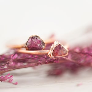 May include: Two rings with raw, deep red gemstones set in gold-colored metal. The rings rest on a bed of purple dried flowers, with a soft, blurred background. The gemstones have a rough, natural texture.