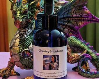 May include: A blue glass bottle with a black spray nozzle and a white label that reads "Cleansing & Protection". The label also includes an image of a woman holding a bowl and a feather. The bottle is in front of a green and purple dragon figurine.