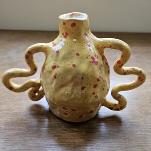 May include: A yellow ceramic vase with red speckles and three handles. The vase has a unique, organic shape.