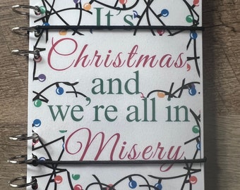 May include: A white spiral bound notebook with a Christmas lights design. The text on the cover reads "It's Christmas and we're all in misery. Save $1,000+."