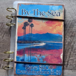 May include: A clear plastic binder with a colorful sunset scene and palm trees. The binder has a brown elastic band and gold rings. The text "By The Sea" is at the top of the image. The text "Save $600 Denominations $1-$9" is at the bottom of the image.