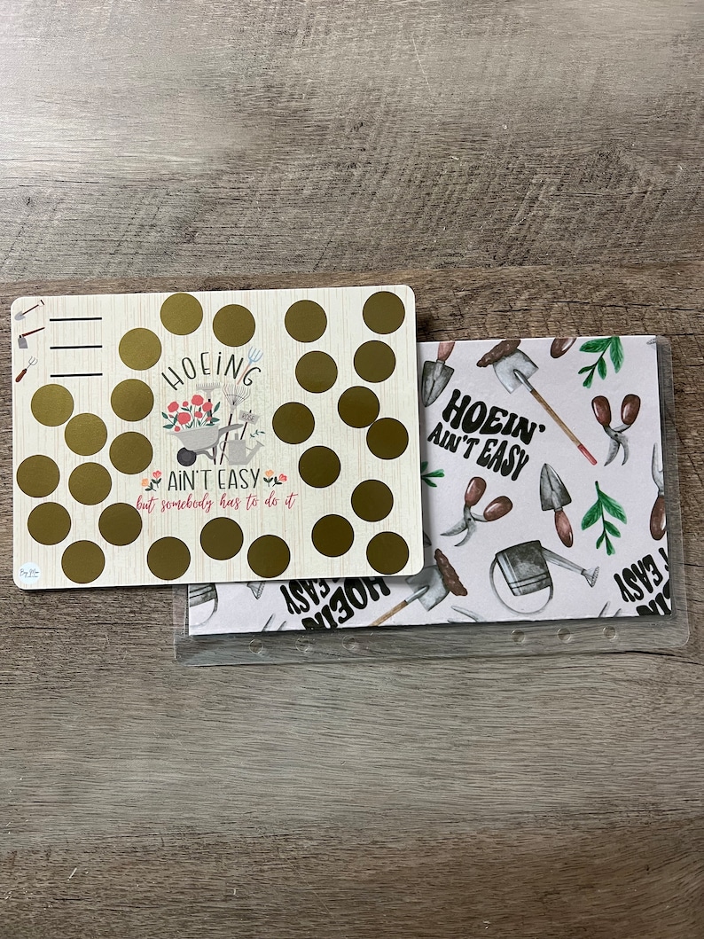 Puede incluir: Una pizarra de borrado en seco dorada y blanca con el texto "HOEING AIN'T EASY but somebody has to do it" y un c&iacute;rculo dorado con una carretilla y flores. La pizarra est&aacute; sobre una l&aacute;mina de pl&aacute;stico transparente con fondo blanco y un patr&oacute;n de herramientas de jardiner&iacute;a y el texto "HOEING AIN'T EASY".