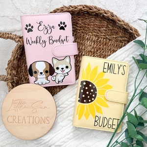 May include: Two personalised budget binders with a cute design. One binder is pink with a dog and puppy illustration and the text "Ezza Weekly Budget". The other binder is yellow with a sunflower illustration and the text "Emily's Budget".