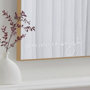 May include: A white framed mirror with the text "you are enough" written in white cursive script. A small white vase with a sprig of burgundy leaves sits in front of the mirror.