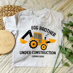 May include: White short-sleeved t-shirt with a yellow and black construction vehicle graphic. The text "Big Brother Under Construction Coming Soon" is printed on the shirt.