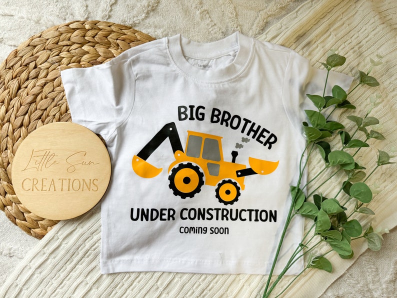 May include: White short-sleeved t-shirt with a yellow and black construction vehicle graphic. The text "Big Brother Under Construction Coming Soon" is printed on the shirt.