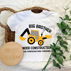 May include: White short-sleeved t-shirt with a yellow construction vehicle graphic and the text "Big Brother Under Construction Expected Completion Date: February 2".