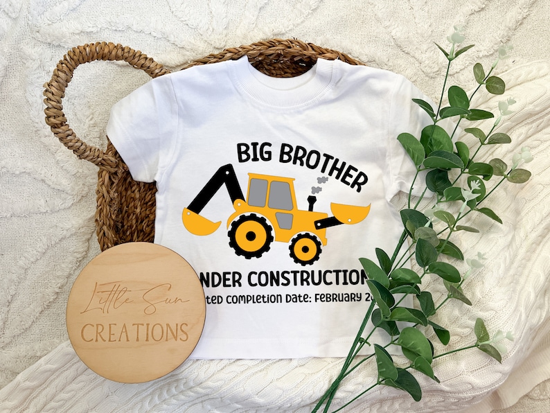 May include: White short-sleeved t-shirt with a yellow construction vehicle graphic and the text "Big Brother Under Construction Expected Completion Date: February 2".