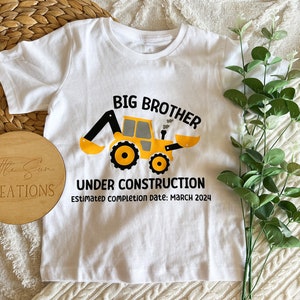 May include: White t-shirt with a yellow construction vehicle graphic and the text "Big Brother Under Construction Estimated Completion Date: March 2024".