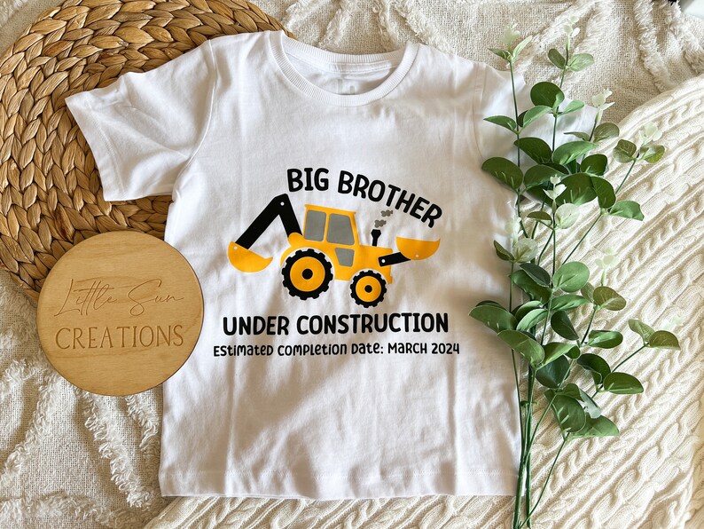 May include: White t-shirt with a yellow construction vehicle graphic and the text "Big Brother Under Construction Estimated Completion Date: March 2024".