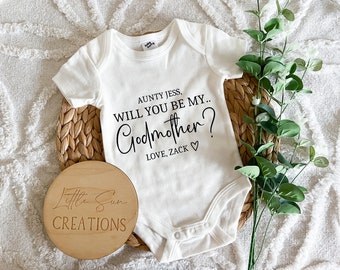 Puede incluir: Un body blanco para bebé con texto negro que dice "AUNTY JESS, WILL YOU BE MY... GODMOTHER? LOVE, ZACK" con un símbolo de corazón. Un letrero redondo de madera con el texto "Little Sun CREATIONS" está en primer plano.