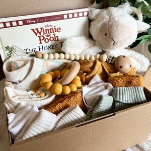 May include: A brown cardboard box filled with baby items, including a Winnie the Pooh book, a plush lamb toy, a wooden bead necklace, a yellow and wooden teether, and several white and mustard yellow baby clothes.