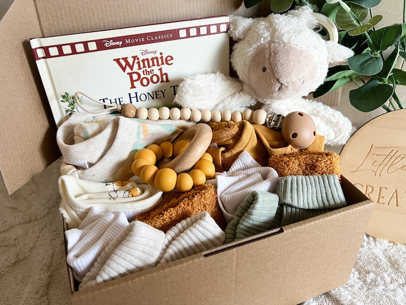 May include: A brown cardboard box filled with baby items, including a Winnie the Pooh book, a plush lamb toy, a wooden bead necklace, a yellow and wooden teether, and several white and mustard yellow baby clothes.