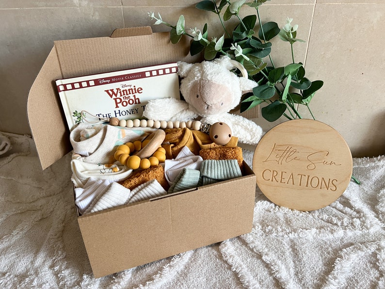 May include: A brown cardboard box filled with baby items, including a book titled "Winnie the Pooh The Honey Tree", a white plush lamb, a wooden teether, and a yellow silicone teether. The box is labelled "Little Sun Creations".