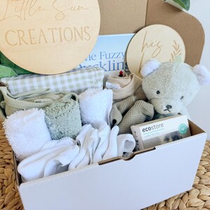 May include: A white gift box filled with baby items, including a green and white gingham blanket, white towels, a green and white striped onesie, a green and white striped hat, a green and white striped stuffed animal, and a small white box with the text "ecostore" on it. The box is decorated with two wooden signs, one that says "Little Sun Creations" and the other that says "Hey Baby".