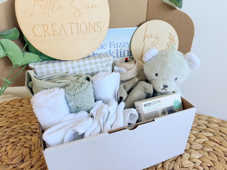 May include: A white gift box filled with baby items, including a green and white gingham blanket, white towels, a green and white striped onesie, a green and white striped hat, a green and white striped stuffed animal, and a small white box with the text "ecostore" on it. The box is decorated with two wooden signs, one that says "Little Sun Creations" and the other that says "Hey Baby".