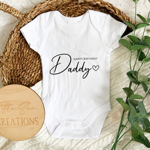 May include: White baby grow with black text that says "Happy Birthday Daddy" with a heart. The grow is on a white knitted surface with a wooden sign that says "Little Sun Creations".