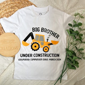 May include: White t-shirt with a yellow construction vehicle graphic and the text "Big Brother Under Construction Estimated completion date: March 2024".