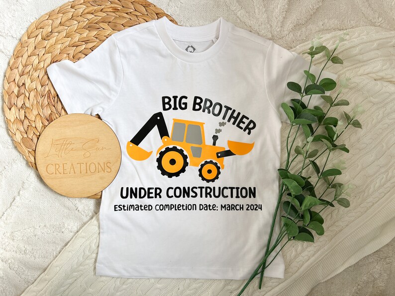 May include: White t-shirt with a yellow construction vehicle graphic and the text "Big Brother Under Construction Estimated completion date: March 2024".