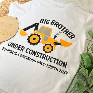 May include: White t-shirt with a yellow construction vehicle graphic and the text "Big Brother Under Construction Estimated completion date: March 2024".