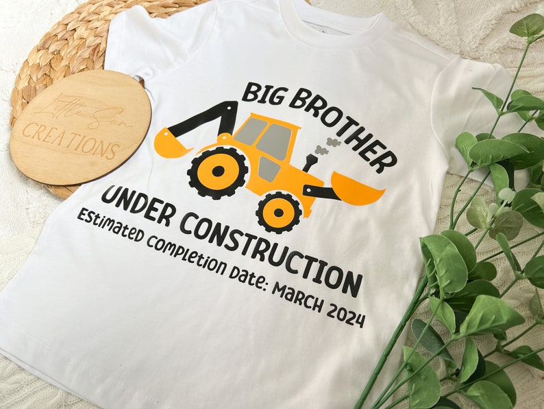 May include: White t-shirt with a yellow construction vehicle graphic and the text "Big Brother Under Construction Estimated completion date: March 2024".