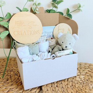 May include: A white gift box filled with baby items, including a plush teddy bear, a book, a wooden sign that says "Little Sun Creations", and other baby essentials. The box is sitting on a woven surface.