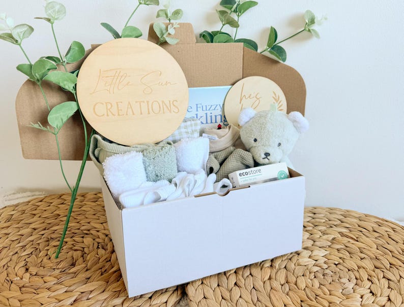 May include: A white gift box filled with baby items, including a plush teddy bear, a book, a wooden sign that says "Little Sun Creations", and other baby essentials. The box is sitting on a woven surface.