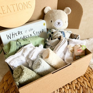 May include: A brown cardboard box filled with baby items, including a green onesie, white socks, a green and white towel, a beige washcloth, a book titled "Where is the Green Sheep?" and a plush grey bear toy.