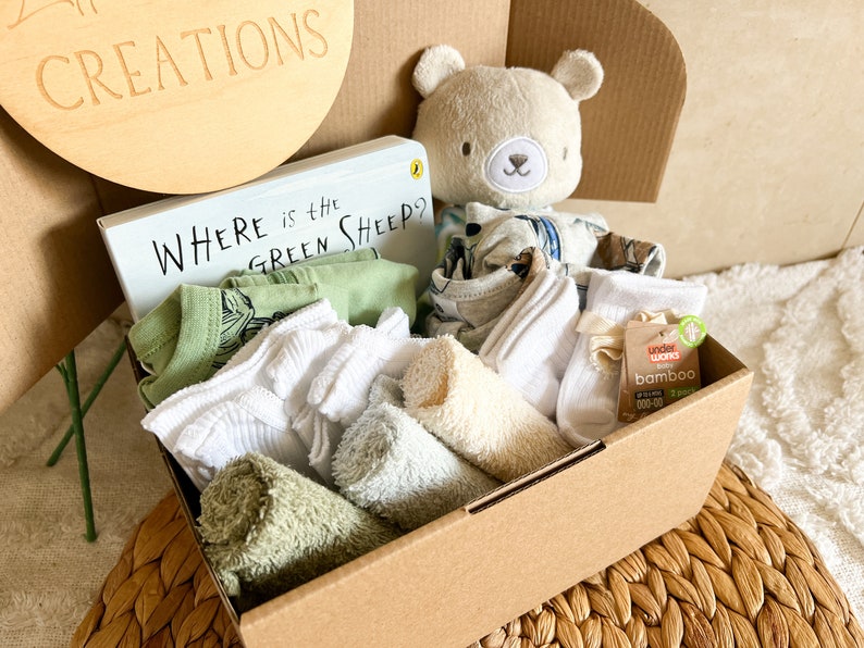 May include: A brown cardboard box filled with baby items, including a green onesie, white socks, a green and white towel, a beige washcloth, a book titled "Where is the Green Sheep?" and a plush grey bear toy.