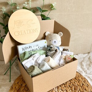May include: A brown cardboard box filled with a baby gift set. The box contains a book titled "Where is the Green Sheep?", a plush teddy bear, a green and white baby onesie, white socks, and green and white washcloths. The box is decorated with a wooden circle that says "Little Sun Creations".