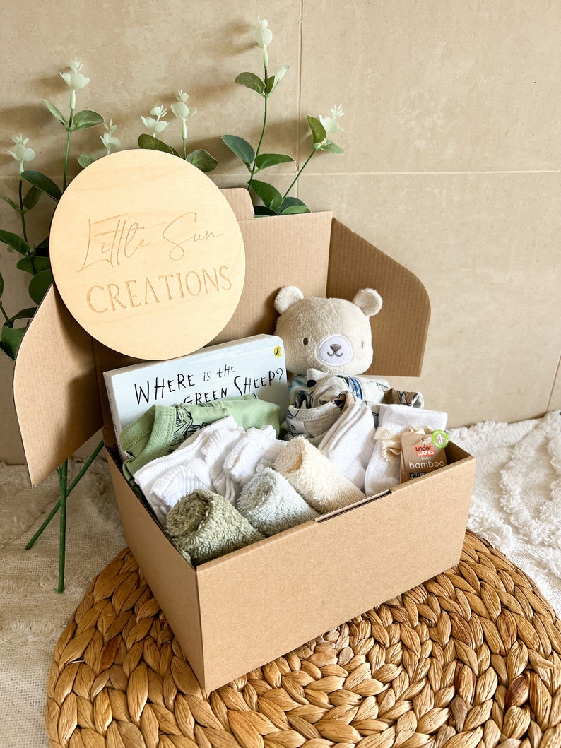 May include: A brown cardboard box filled with a baby gift set. The box contains a book titled "Where is the Green Sheep?", a plush teddy bear, a green and white baby onesie, white socks, and green and white washcloths. The box is decorated with a wooden circle that says "Little Sun Creations".