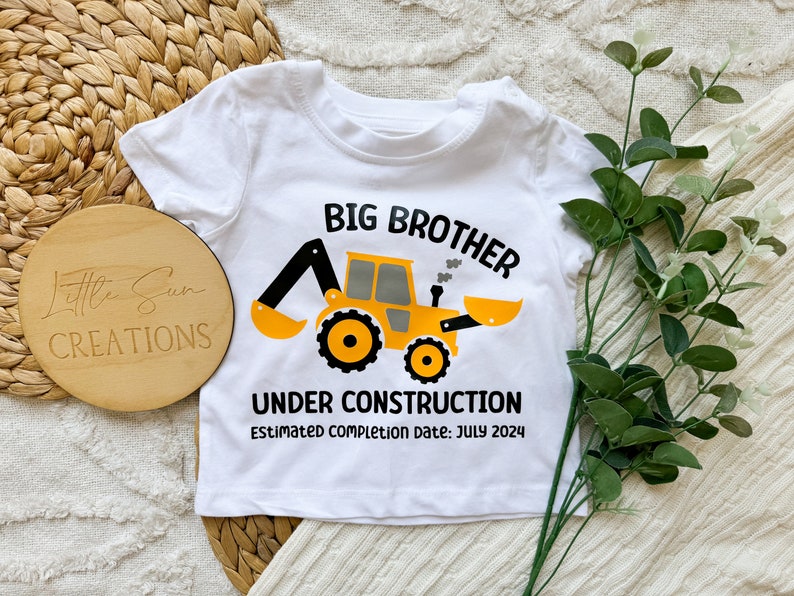 May include: White baby onesie with a yellow construction vehicle graphic and the text "Big Brother Under Construction Estimated completion date: July 2024".