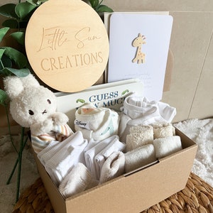 May include: A brown cardboard box filled with baby clothes and accessories. The box is decorated with a wooden sign that says "Little Sun Creations". The box also contains a stuffed animal, a book, and a card.