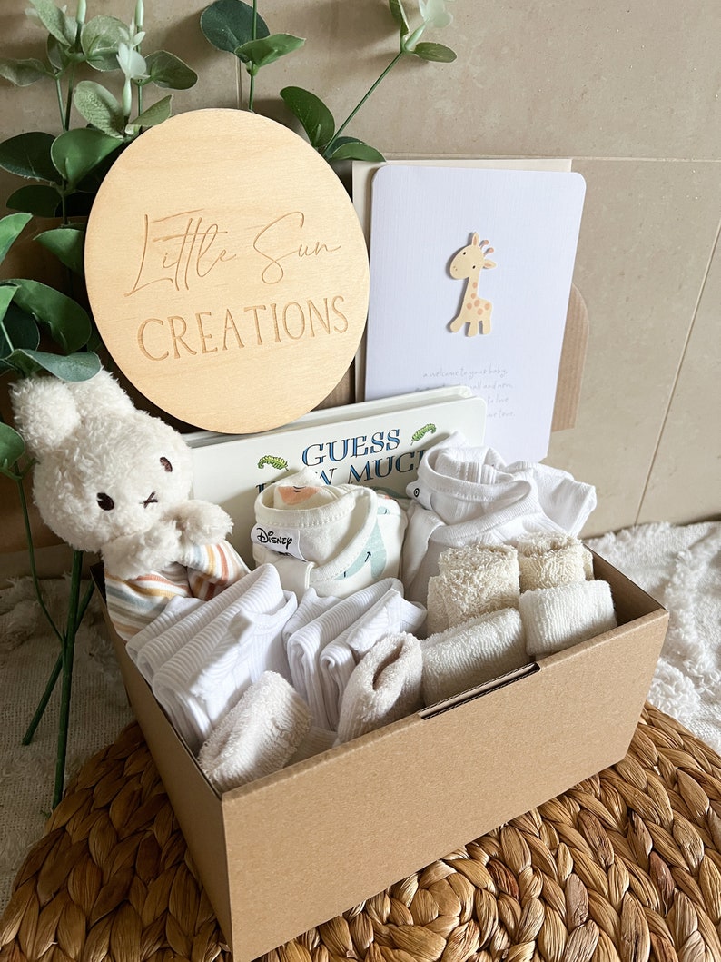 May include: A brown cardboard box filled with baby clothes and accessories. The box is decorated with a wooden sign that says "Little Sun Creations". The box also contains a stuffed animal, a book, and a card.