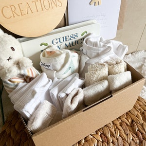 May include: A brown cardboard box filled with baby items, including a white stuffed bunny, a book titled "Guess How Much I Love You", white vests, white socks, and white face cloths.