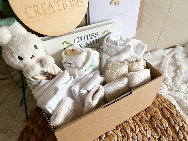 May include: A brown cardboard box filled with baby items, including a white stuffed bunny, a book titled "Guess How Much I Love You", white vests, white socks, and white face cloths.