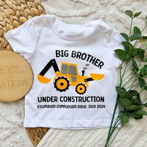 May include: White baby onesie with a yellow construction vehicle graphic and the text "Big Brother Under Construction Estimated completion date: July 2024".